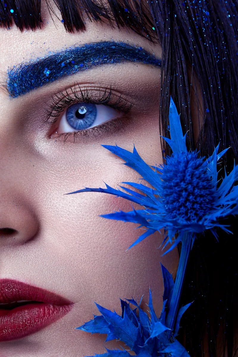 Close-up beauty portrait retouching showing a model with blue glitter eyebrows, red lips and a blue thistle flower