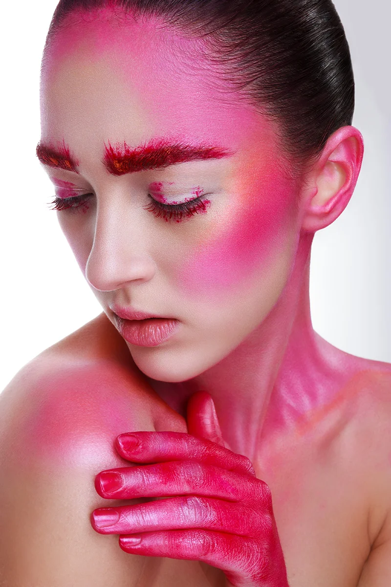Creative beauty portrait retouching showing a model with vibrant pink face and body paint and red glittery eyebrows