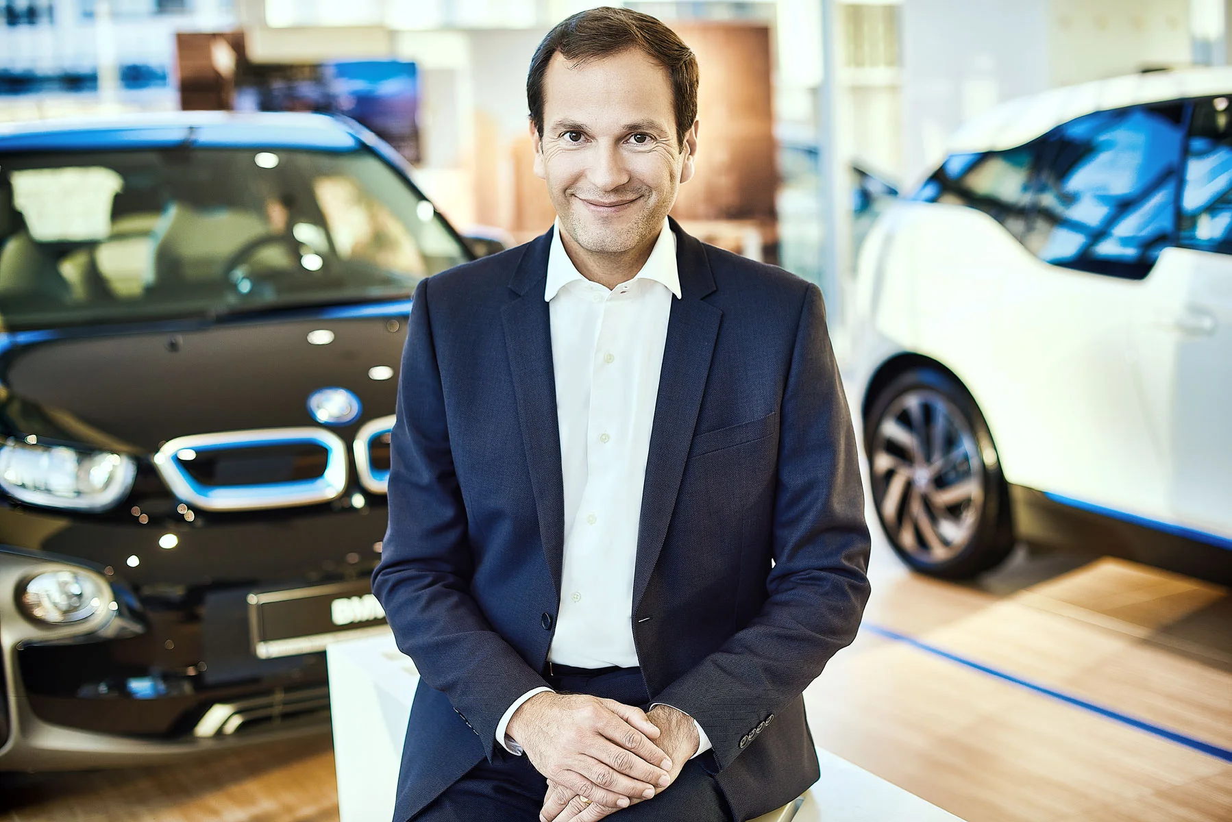 Business portrait retouching of a professional manager sitting in a modern car dealership showroom with electric cars in the background