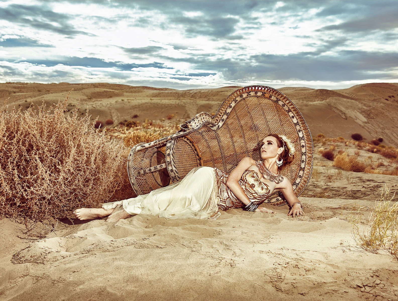 Fashion editorial retouching of a model in a gold dress lying on a large wicker chair in the sand dunes