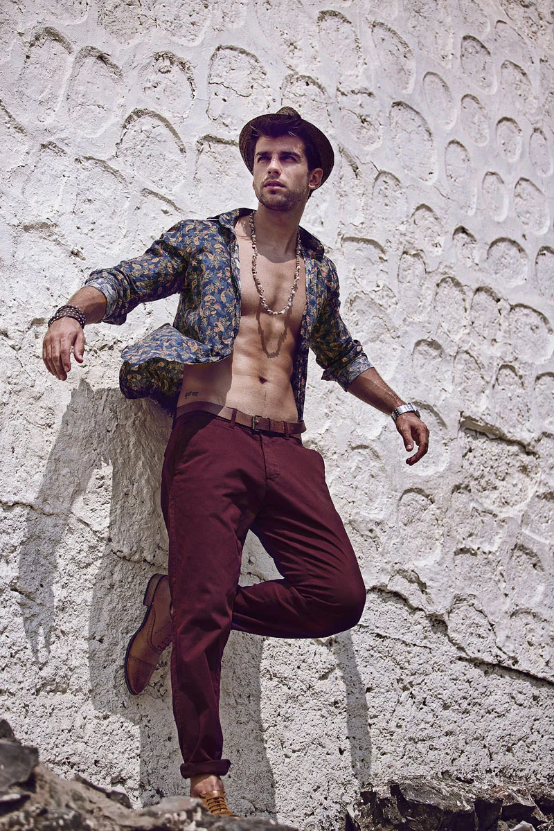 Outdoor fashion photography retouching of a male model in a hat and open paisley shirt leaning against a textured white wall